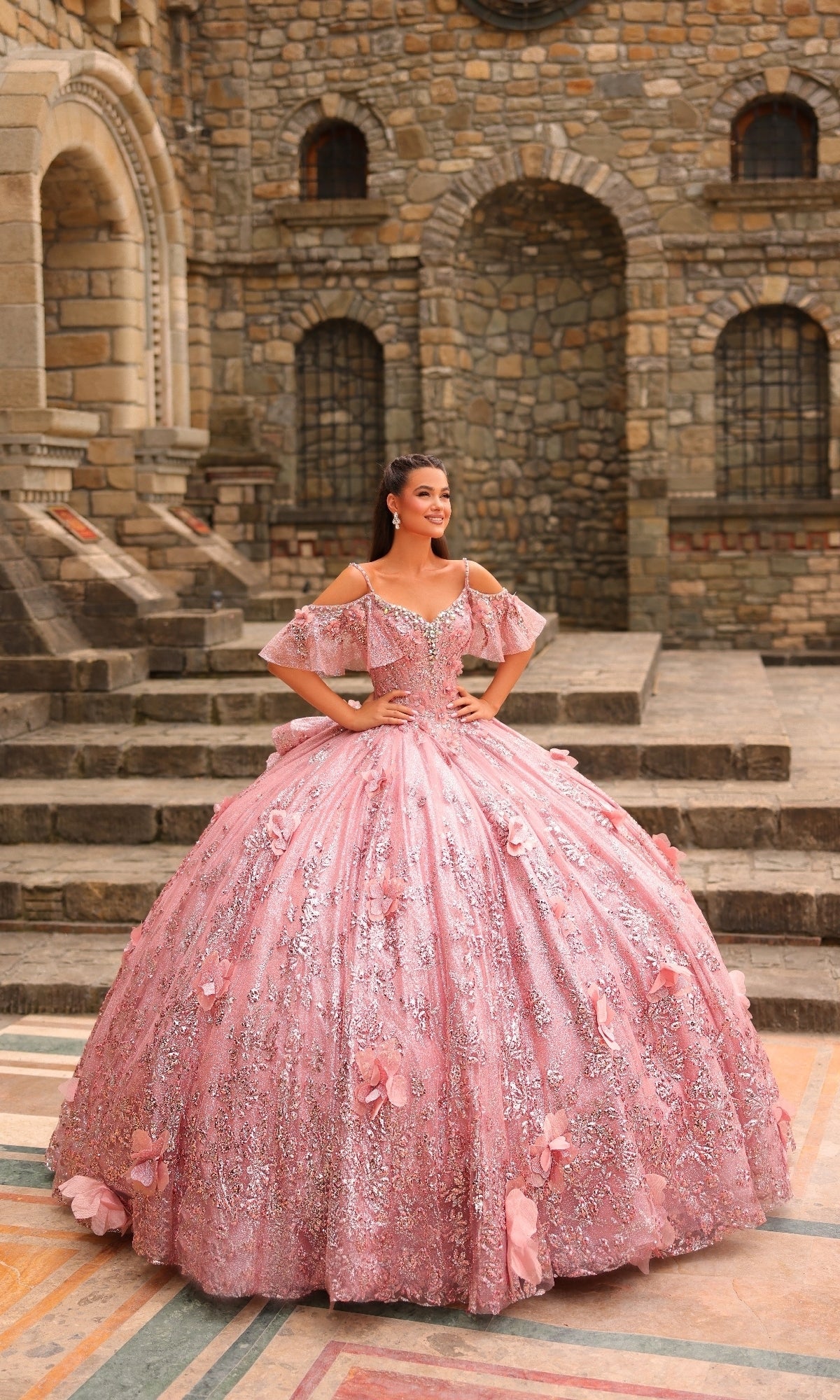 A young woman stands outdoors on stone steps, wearing the Amarra 54229 Glitter Tulle Quinceañera Dress, an elaborate pink ball gown adorned with floral embellishments, beading, and sparkling glitter tulle.